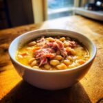 A close-up of a white bowl filled with thick navy bean soup topped with shredded ham, sitting on a rustic wooden table.