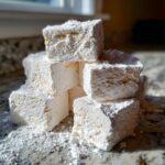 A stack of large, fluffy homemade marshmallows heavily dusted with powdered sugar on a marble counter.