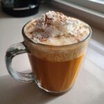 A close-up of a homemade pumpkin spice latte in a clear glass mug, topped with whipped cream and cinnamon.