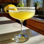 A bright yellow, chilled lemon drop martini in a coupe glass with a sugar rim and lemon twist garnish.