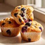 A stack of three moist blueberry protein muffins, one broken open to show the soft interior.
