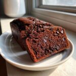 Two slices of moist chocolate bread loaded with chocolate chips, served on a white plate near a window.