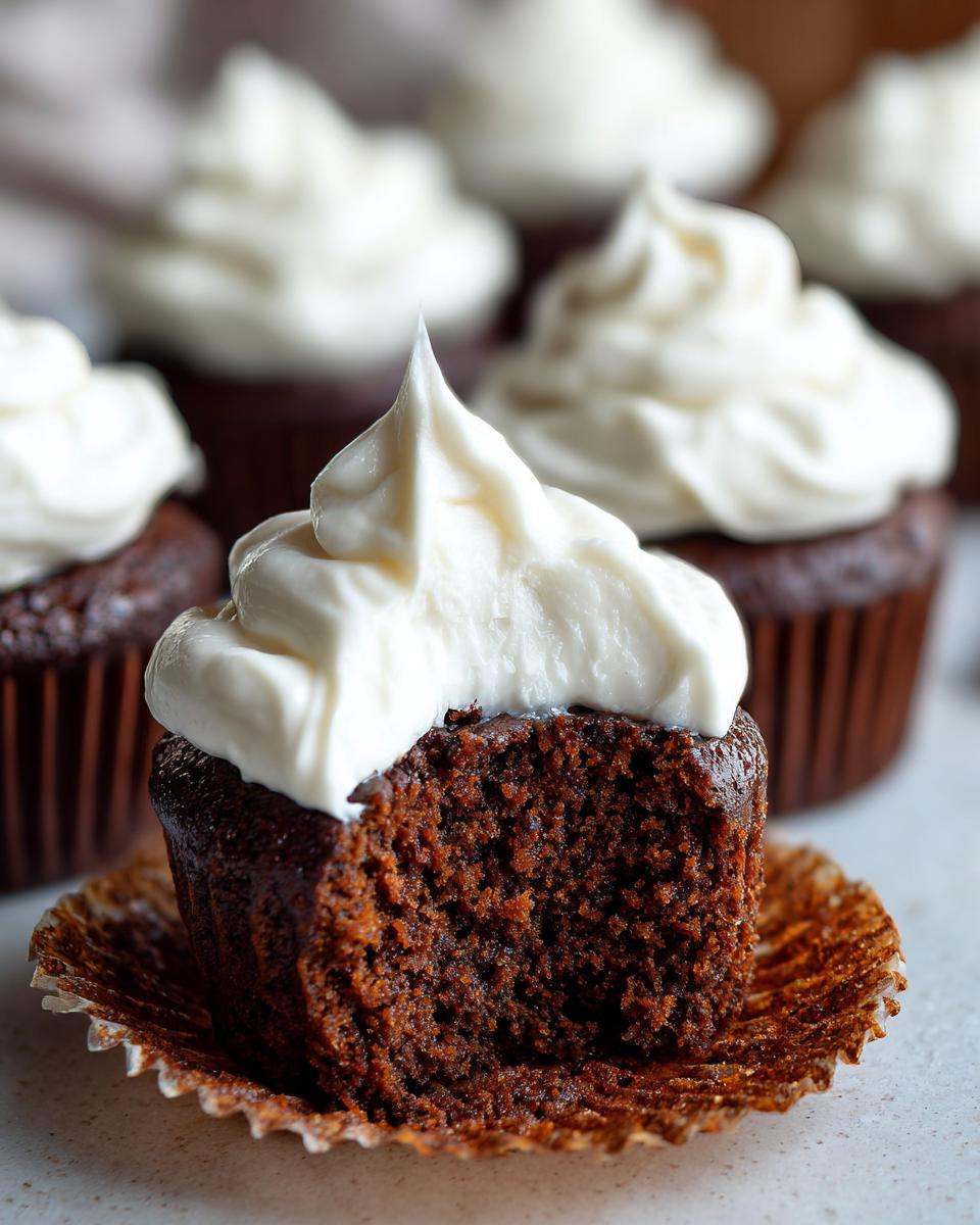 Close-up of a gingerbread cupcakes with a bite taken out, showing the moist texture and topped with white frosting.