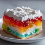 A moist slice of rainbow jello poke cake topped with whipped cream, showing vibrant layered gelatin.