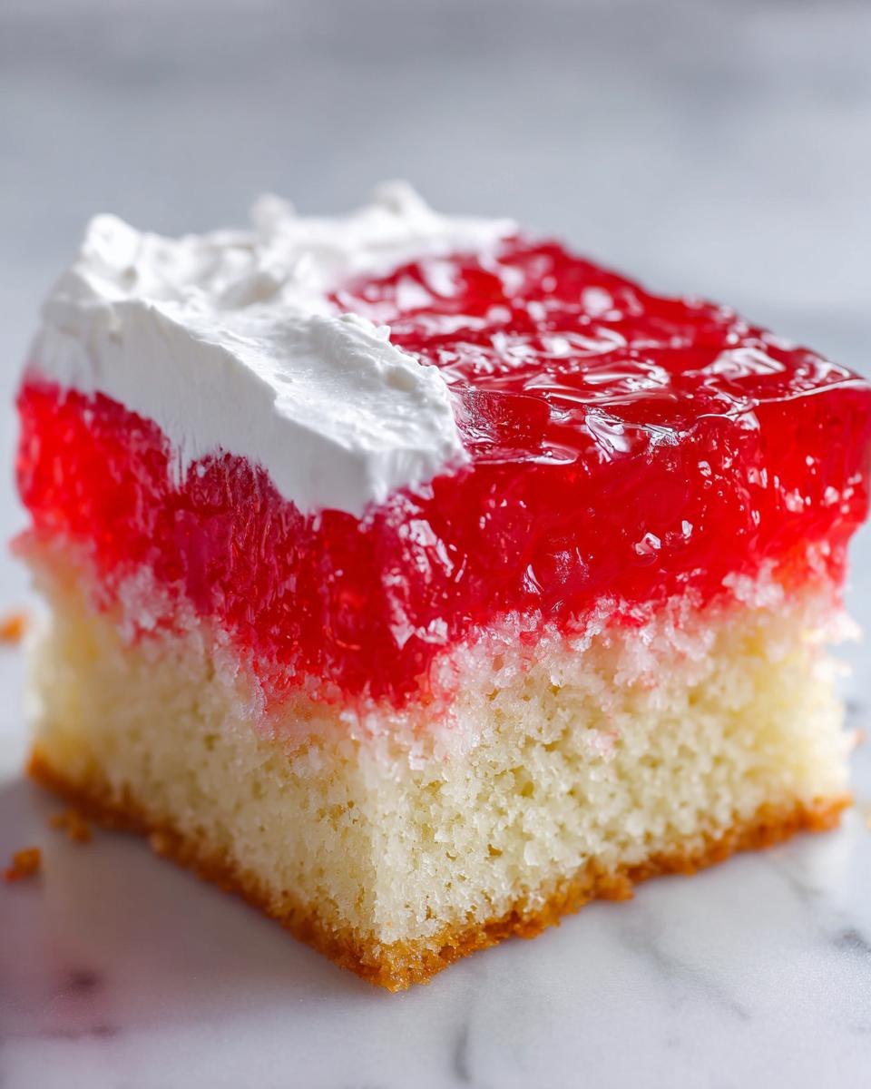 A close-up of a square slice of jello poke cake showing a white cake base, bright red gelatin layer, and white whipped topping.