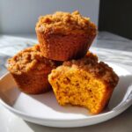 A stack of three moist sweet potato muffins, one with a bite taken out showing the orange interior and crumb topping.
