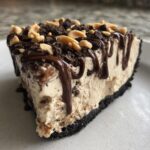 A decadent slice of no-bake ice cream pie with a dark crust, peanut butter filling, chocolate drizzle, and chopped peanuts.
