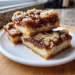Three rich pecan pie bars stacked on a white plate, showing the thick, gooey filling and buttery shortbread crust.
