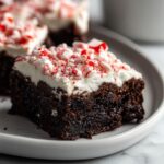 A close-up of a rich, dark chocolate peppermint brownies slice topped with white frosting and crushed candy canes.