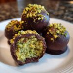 Close-up of several rich Dubai chocolate balls dipped in dark chocolate and topped with crushed pistachios, one is bitten open.