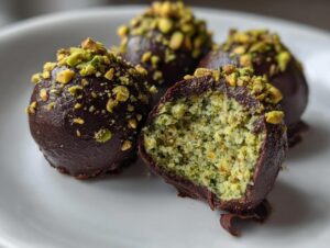 Close-up of rich dark chocolate dubai chocolate balls topped with crushed pistachios, one is cut open showing the green pistachio center.