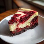 A perfect slice of swirled red velvet cheesecake sits on a white plate, showing the dark crust and creamy texture.