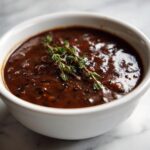A close-up of rich, dark homemade brown gravy garnished with fresh thyme sprigs in a white bowl.