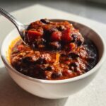 A spoonful of thick, rich black bean chili being lifted from a white bowl, showing chunks of tomato and beans.