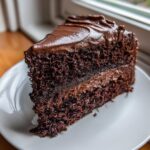 A tempting slice of moist chocolate fudge cake layered with dark frosting, sitting on a white plate.