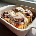 Four gooey small batch cinnamon rolls covered in thick white icing in a white baking dish.
