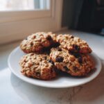 A stack of soft oatmeal raisin cookies piled on a white plate near a sunlit window.