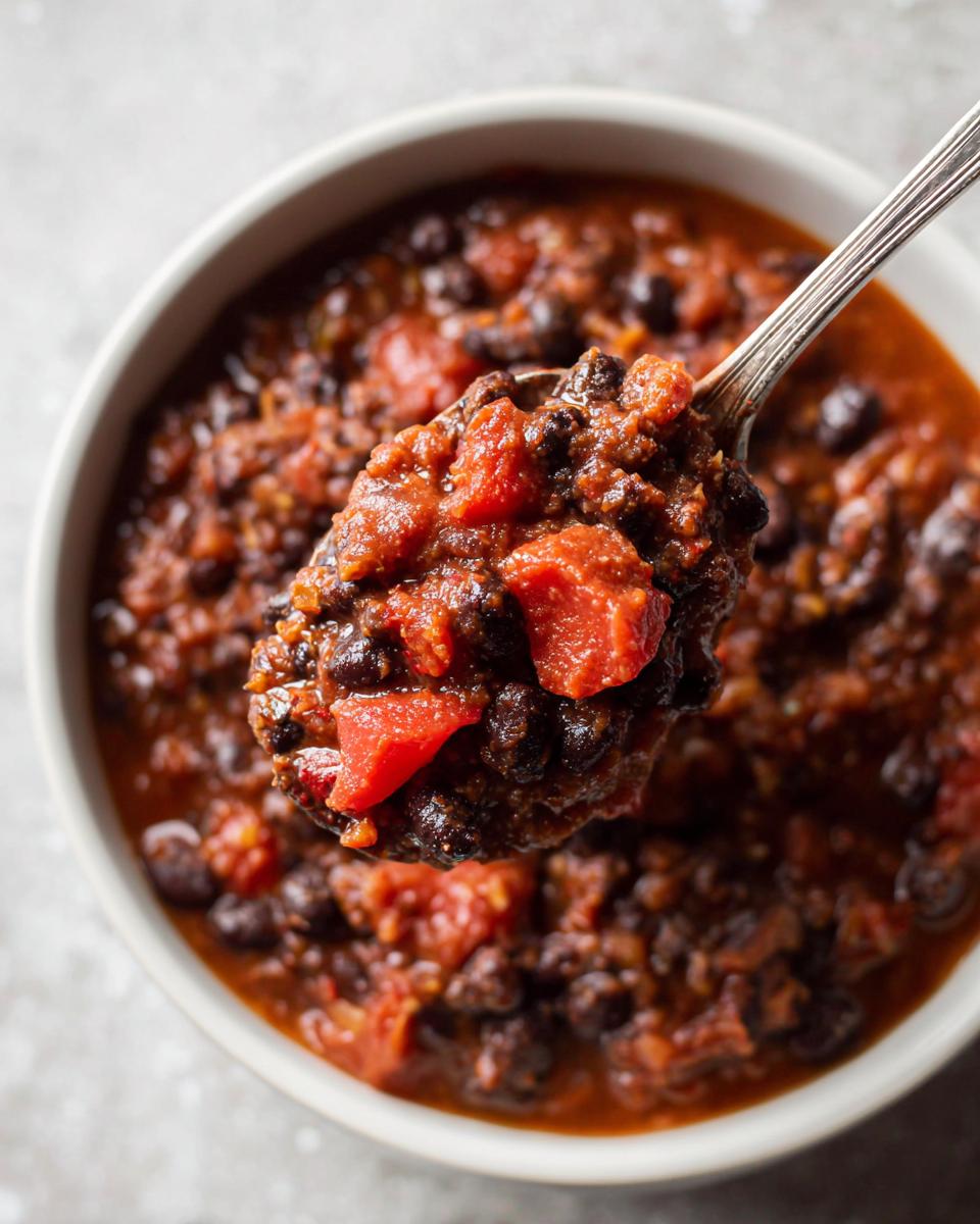 A spoonful of thick, rich black bean chili with visible chunks of tomato being lifted from a bowl.