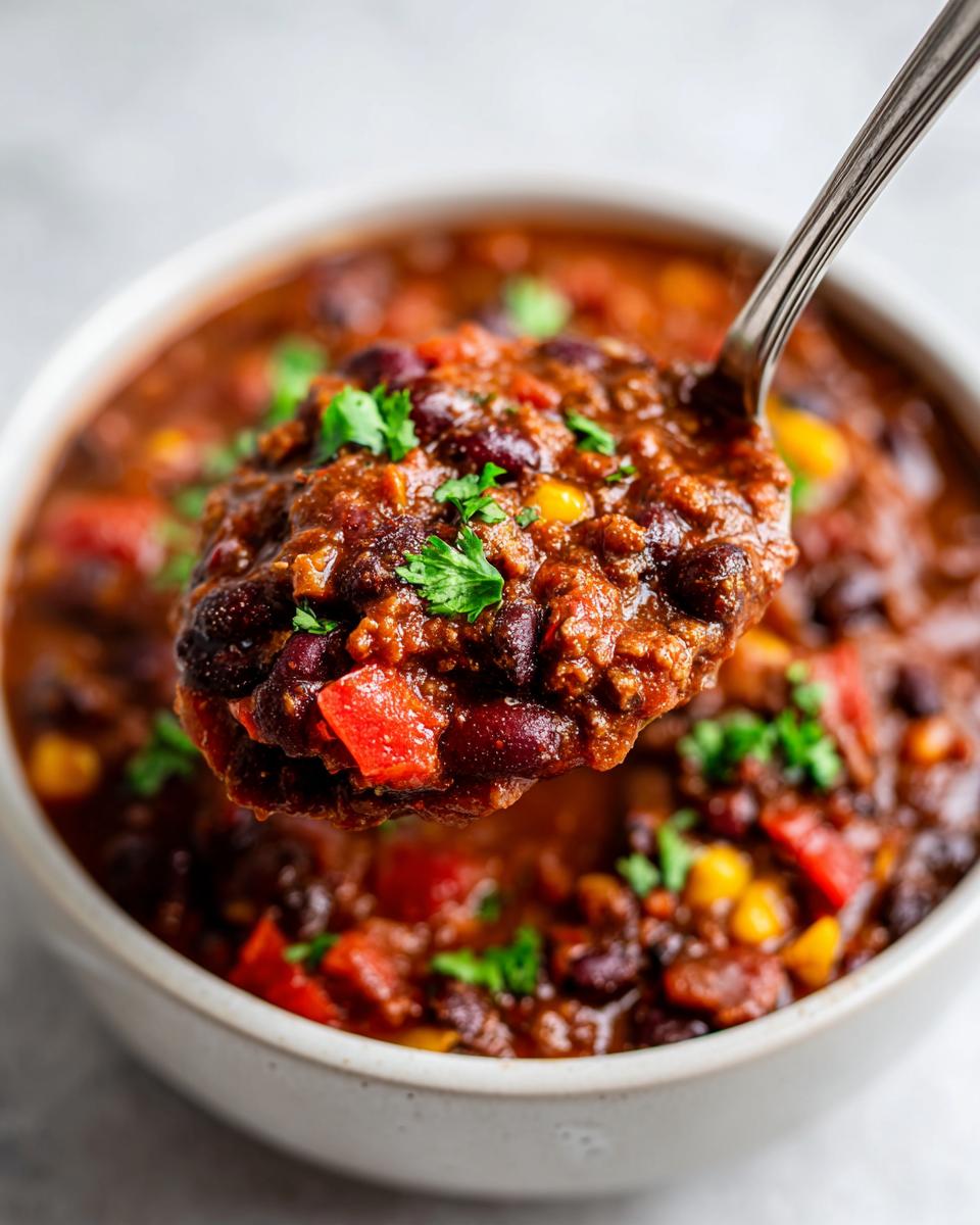 A spoonful of thick, hearty black bean chili, rich with beans and red peppers, being lifted from a white bowl.