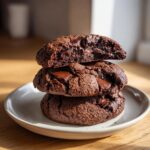 A stack of three rich, dark vegan chocolate chip cookies with melted chocolate chunks on a small plate.