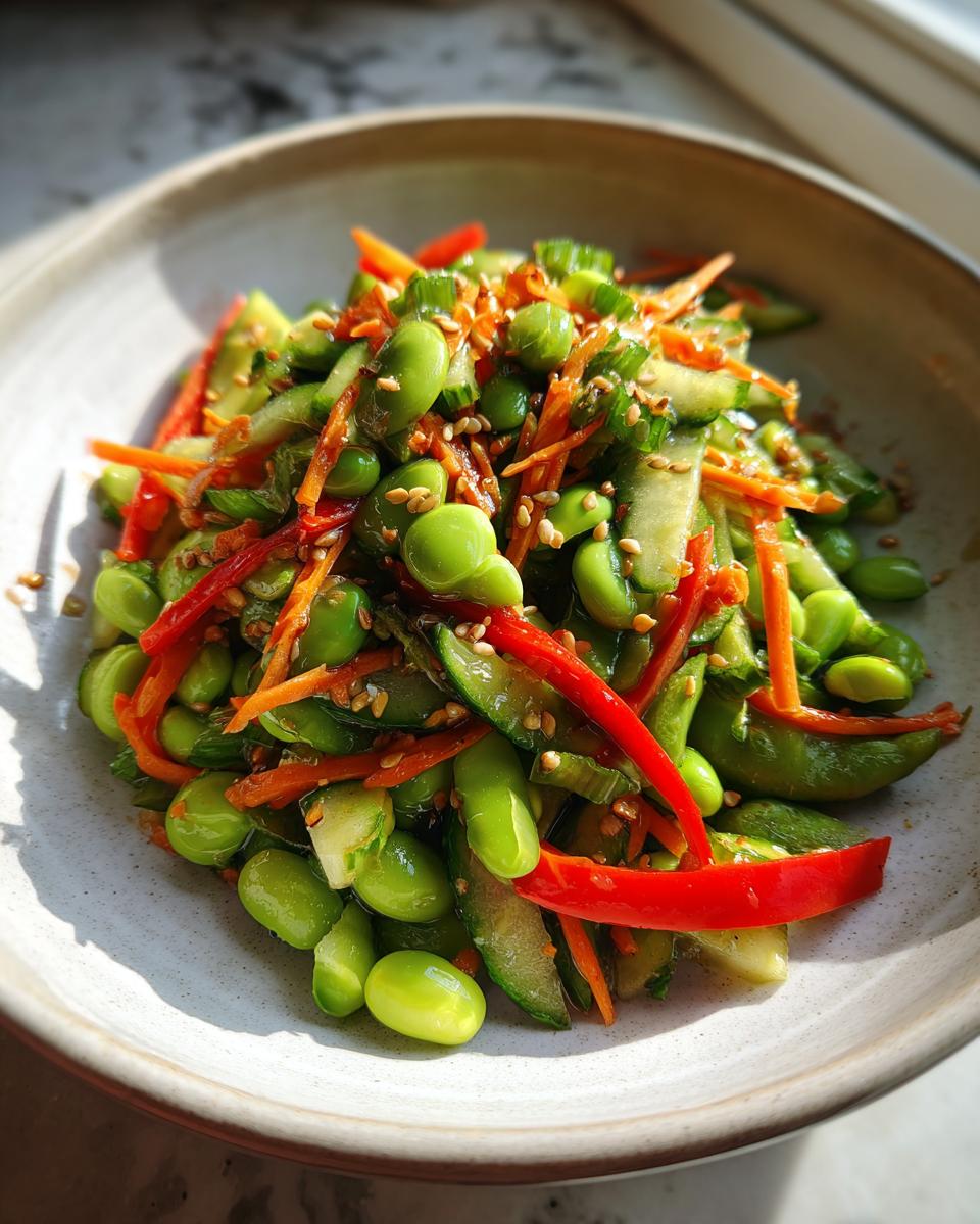 Close-up of a vibrant edamame salad featuring bright green edamame, shredded carrots, sliced cucumber, and red pepper strips, topped with sesame seeds.