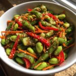A close-up of a vibrant edamame salad featuring shelled edamame, shredded carrots, cucumber chunks, and sliced red peppers, tossed in a sesame dressing.