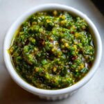 Close-up of thick, chunky green tomato salsa with visible chili flakes served in a small white bowl.