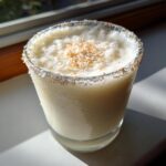 A creamy white Christmas margarita in a glass with a coconut-rimmed edge and toasted coconut flakes on top.