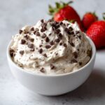 A white bowl filled with creamy, light-colored chocolate dip topped with mini chocolate chips, with strawberries in the background.