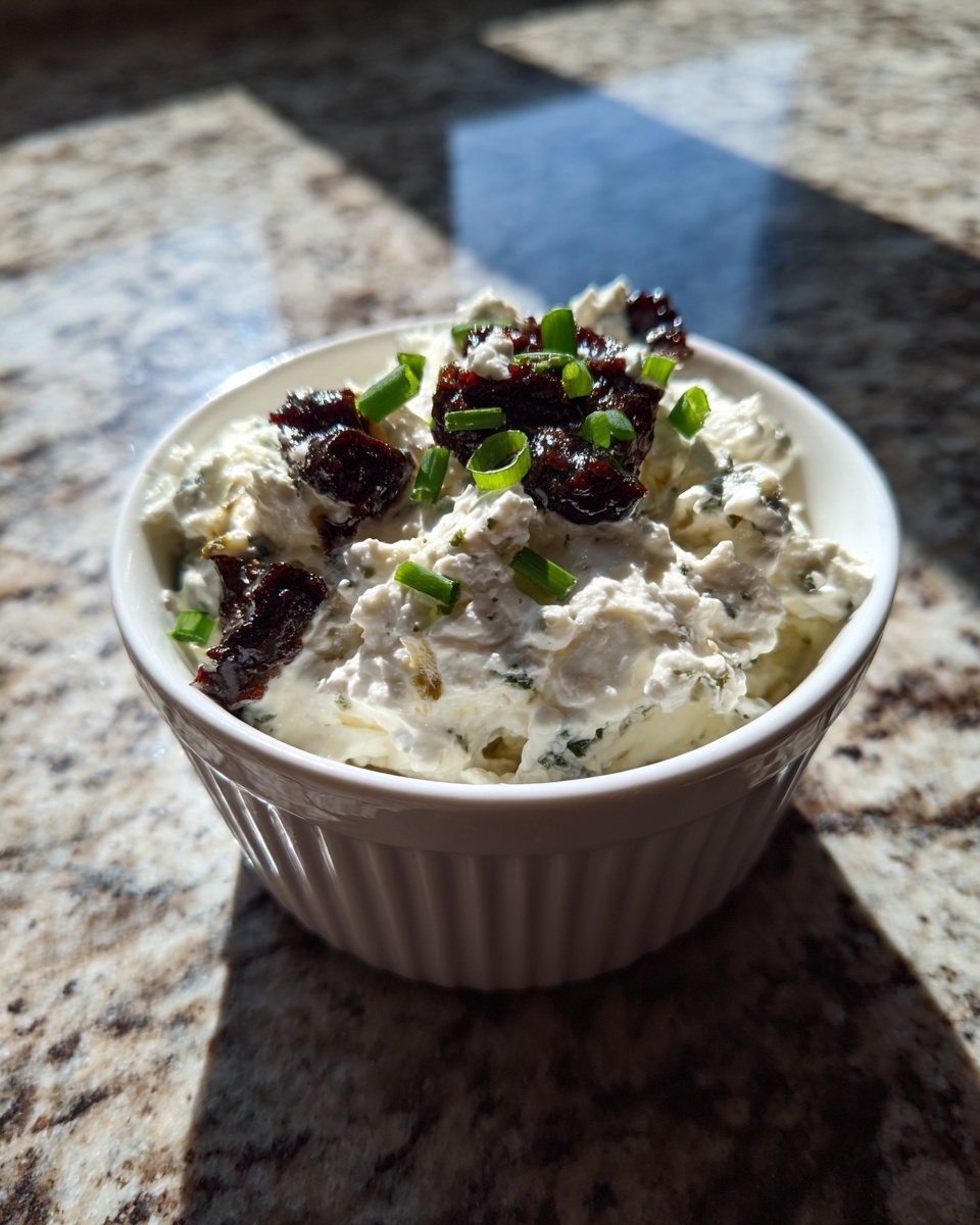 A small white bowl filled with creamy blue cheese dip, topped with dark sun-dried tomatoes and chopped green onions.