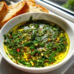 Close-up of a white bowl filled with flavorful olive oil bread dip, featuring fresh green herbs and red chili flakes.