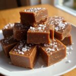 A stack of rich, brown squares of homemade salted caramel fudge topped generously with flaky sea salt.