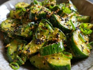 Close-up of sliced cucumbers coated in a creamy peanut butter dressing, topped with black and white sesame seeds and green onions, for an asian cucumber salad.
