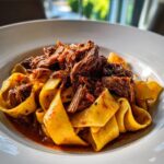Close-up of rich braised short rib pappardelle pasta topped with shredded beef in a white bowl.