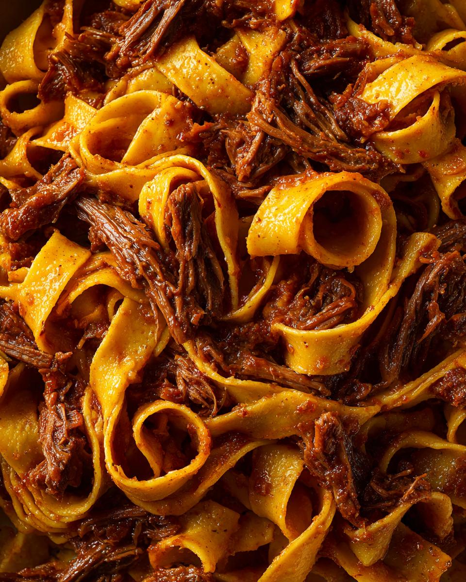 A rich, close-up view of tender shredded braised short rib pappardelle pasta coated in a deep red sauce.