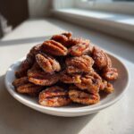 A close-up of a pile of glistening candied pecans for salad, coated in sugar crystals, sitting on a white dish.