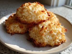 A close-up of three golden-brown, chewy coconut cookies stacked on a light gray plate.
