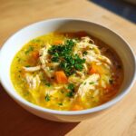 Close-up of a white bowl filled with bright yellow chicken orzo soup, topped with shredded chicken, carrots, and fresh parsley.