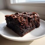 A close-up of a rich, fudgy chocolate date brownies slice topped with melted chocolate chips on a white plate.