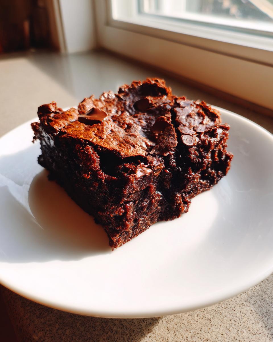 A fudgy square of chocolate date brownies sits on a white plate, catching sunlight near a window.