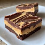Two squares of rich chocolate peanut butter fudge with a marbled chocolate top layer, served on a white plate.