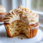 A close-up of a churro cupcake with a bite taken out, showing the moist cake texture and cinnamon-dusted frosting.