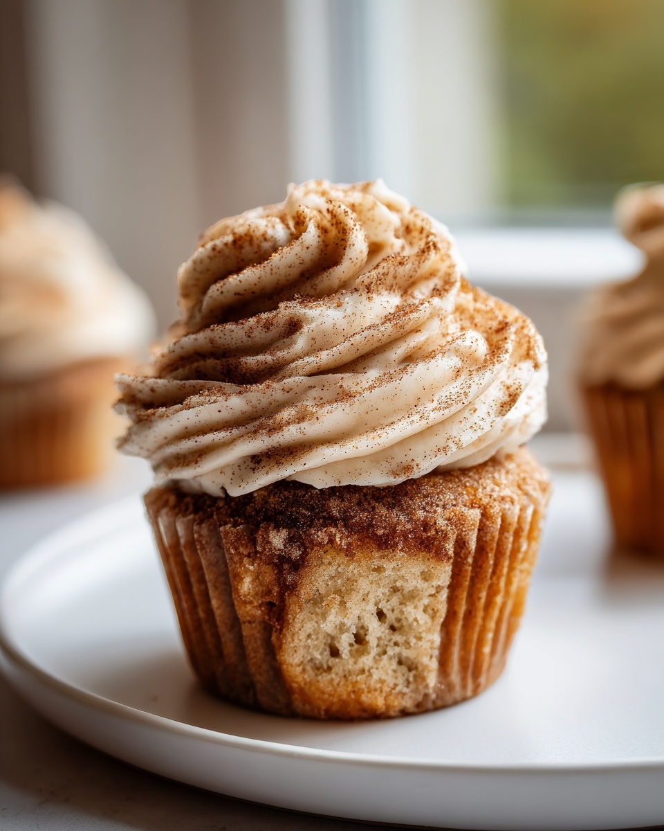 A close-up of a single churro cupcake topped with swirled cinnamon frosting, sitting on a white plate.