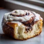 A close-up shot of a single, fluffy cinnamon rolls homemade pastry topped generously with white cream cheese frosting.