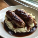 Two browned sausages served over creamy mashed potatoes, covered in dark gravy, classic bangers and mash.