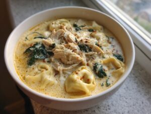 Close-up of a bowl of creamy chicken tortellini soup topped with shredded chicken, spinach, and red pepper flakes.