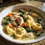 Close-up of a white bowl filled with creamy chicken tortellini soup, featuring chunks of chicken, spinach, and carrots.