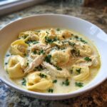 A close-up of a white bowl filled with creamy chicken tortellini soup crock pot, topped with shredded chicken and parsley.