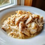 A close-up of shredded creamy garlic chicken crockpot served in a white bowl near a window.