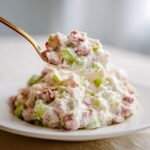 A spoonful of creamy ham salad recipe, featuring diced ham and celery, being lifted from a mound on a white plate.